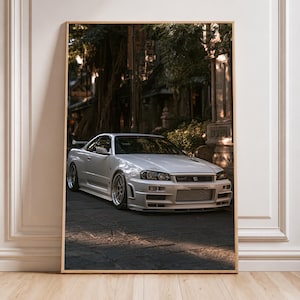 May include: A framed vertical wall art print featuring a white Nissan Skyline GT-R R34 sports car parked on a paved street. The car has gold-colored wheels, a front bumper with a large air intake, and a rear spoiler, set against a backdrop of trees.