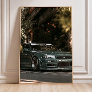 Könnte beinhalten: Ein gerahmter vertikaler Wandkunstdruck, der einen dunkel salbeigrünen Nissan Skyline GT-R R34 Sportwagen auf einer Asphaltstraße zeigt. Das Fahrzeug verfügt über eine tiefergelegte Federung, bronzefarbene Felgen und einen Frontstoßfänger.