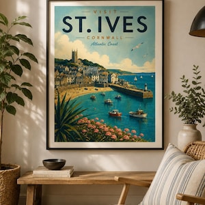 May include: A framed travel poster titled VISIT ST. IVES CORNWALL Atlantic Coast. The illustration depicts a coastal town with a stone church on a hill, a sandy beach, a harbor with small boats, and pink flowers in the foreground under a blue sky.