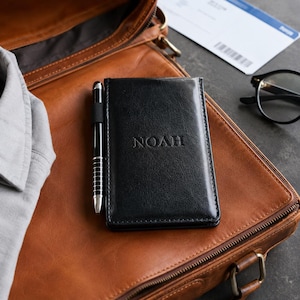 以下が含まれることがあります： フロントカバーにNOAHの名前が刻印された黒いレザー製のパスポートホルダー。ホルダーのサイドループには、洗練された黒いペンが固定されています。このアイテムは滑らかで光沢のある仕上げが特徴で、縁にはきれいなステッチが施されています。