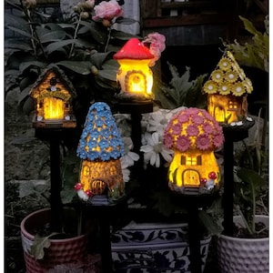 Könnte beinhalten: Fünf beleuchtete Gartenstecker in Form von Miniaturhäusern und Pilzen. Die Designs umfassen ein braunes Haus mit Welcome-Schild, einen roten Pilz, ein blaues blumenbedecktes Haus, ein rosa blumenbedecktes Haus und ein gelbes Haus mit Blütendach.