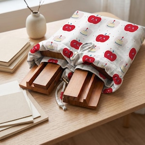 May include: A white fabric drawstring bag featuring a repeating pattern of red cartoon apples with faces and small birthday cakes with candles. Inside the bag are two rectangular wooden stands with grooved surfaces for holding items.