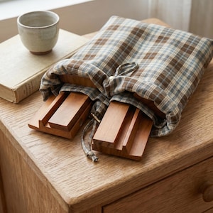 May include: A brown and blue plaid fabric drawstring pouch containing two rectangular wooden stands. The pouch features a gathered top with a matching fabric cord and a small metallic bead closure, partially revealing the smooth wooden surfaces.