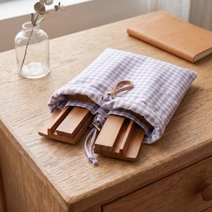 May include: A light purple and white gingham fabric drawstring pouch containing two rectangular wooden stands. The pouch features a small tan leather loop at the top and white cord strings at the bottom, partially revealing the natural wood grain.
