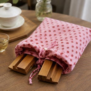 May include: A pink fabric drawstring pouch featuring a repeating pattern of small red strawberries. The pouch is partially open, revealing two rectangular wooden stands with grooved slots inside. The bag is secured with a matching pink drawstring cord.