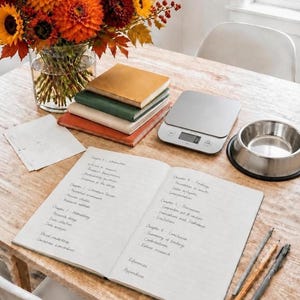 May include: An open notebook with handwritten chapter outlines, a stack of three books, a digital scale, a metal bowl, and a glass vase filled with orange and red autumn flowers. Three pens lie on the wooden surface next to the notebook.