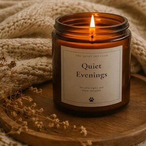 K&ouml;nnte beinhalten: Eine brennende Kerze in einem zylindrischen bernsteinfarbenen Glasgef&auml;&szlig; mit einem wei&szlig;en Etikett, das THE QUIET PET CLUB, Quiet Evenings, for slow nights and warm company sowie ein kleines Pfotenabdruck-Symbol zeigt. Die Kerze hat eine Flamme.