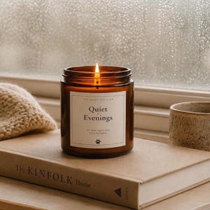 K&ouml;nnte beinhalten: Eine brennende Kerze in einem zylindrischen bernsteinfarbenen Glasgef&auml;&szlig; mit einem wei&szlig;en Etikett, das The Quiet Pet Club Quiet Evenings for slow nights and warm company und ein Pfotenabdruck-Symbol zeigt. Die Kerze steht auf einem beigen Buch.