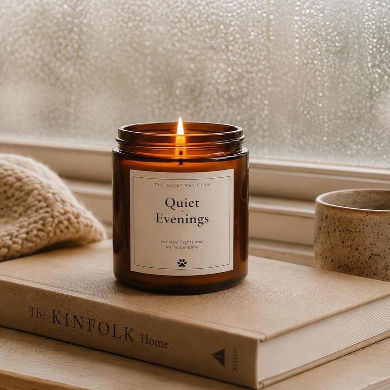 K&ouml;nnte beinhalten: Eine brennende Kerze in einem zylindrischen bernsteinfarbenen Glasgef&auml;&szlig; mit einem wei&szlig;en Etikett, das The Quiet Pet Club Quiet Evenings for slow nights and warm company und ein Pfotenabdruck-Symbol zeigt. Die Kerze steht auf einem beigen Buch.