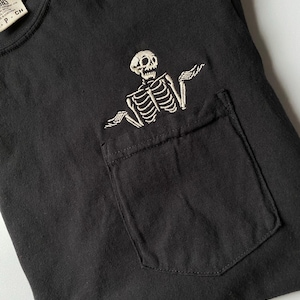 May include: A black t-shirt featuring a white embroidered skeleton graphic positioned above a chest pocket. The skeleton is depicted with arms raised in a shrug gesture. A partial clothing label is visible at the neckline with text reading CH.
