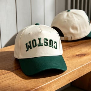 Puede incluir: Una gorra de béisbol color crema con visera verde oscuro y la palabra CUSTOM bordada en letras arqueadas de color verde oscuro en el panel frontal. Una segunda gorra a juego es parcialmente visible al fondo con un cierre ajustable.