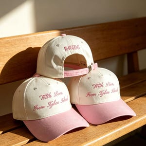 May include: Three cream-colored baseball caps with pink brims and pink embroidery. Two caps feature the text "With Love, From Tybee Island" in cursive script, while the top cap displays the word "BRIDE" in pink lettering above the adjustable strap.