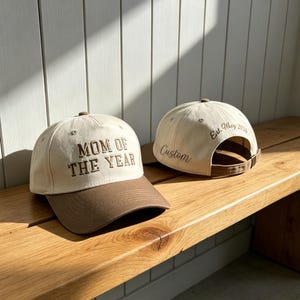 Puede incluir: Dos gorras de b&eacute;isbol con corona color crema y visera marr&oacute;n. La gorra delantera muestra el texto bordado MOM OF THE YEAR. La gorra trasera presenta el texto bordado Est. May 2026 y la palabra Custom en una fuente de escritura cursiva.