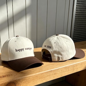 May include: Two baseball caps with off-white crowns and dark brown brims. The front cap features the embroidered text "happy camper" in dark brown. The back cap shows the rear view with an adjustable strap and the embroidered text "est. 2026".