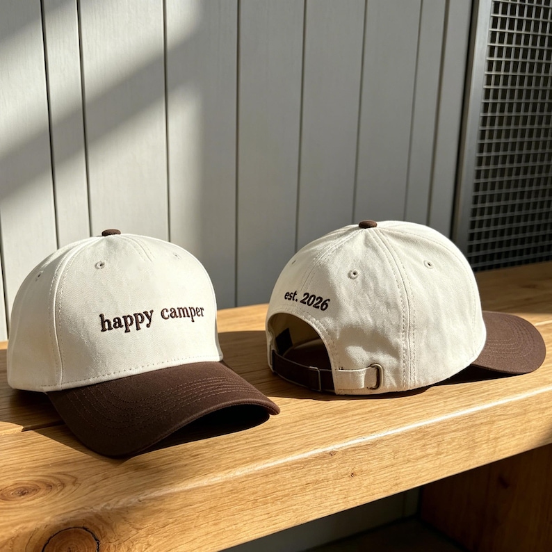 May include: Two baseball caps with off-white crowns and dark brown brims. The front cap features the embroidered text "happy camper" in dark brown. The back cap shows the rear view with an adjustable strap and the embroidered text "est. 2026".