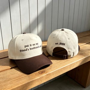 May include: Two baseball caps featuring a cream-colored crown and a dark brown brim. The front cap displays the text "put it on my friend's husband's tab" in dark brown embroidery, while the back cap shows "est. 2026" with an adjustable strap.