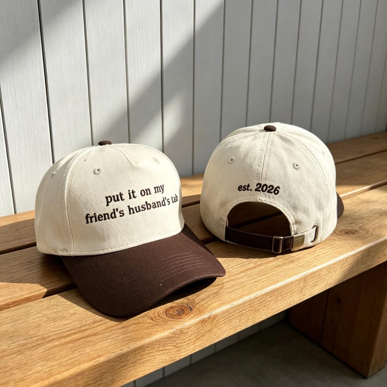 May include: Two baseball caps featuring a cream-colored crown and a dark brown brim. The front cap displays the text "put it on my friend's husband's tab" in dark brown embroidery, while the back cap shows "est. 2026" with an adjustable strap.