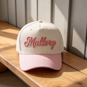Puede incluir: Una gorra de béisbol de color crema con el nombre Mallory bordado en letras cursivas rosas en relieve en el panel frontal. La gorra tiene una visera rosa sólida con costuras visibles y una corona estructurada con ojales para ventilación.