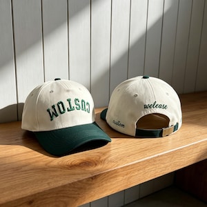 Könnte beinhalten: Zwei Baseballkappen mit cremefarbenen Kronen und dunkelgrünen Schirmen. Die vordere Kappe zeigt das Wort CUSTOM in dunkelgrünen Stickbuchstaben. Die hintere Kappe zeigt das Wort welease in grüner Schreibschrift und custom am seitlichen Riemen.