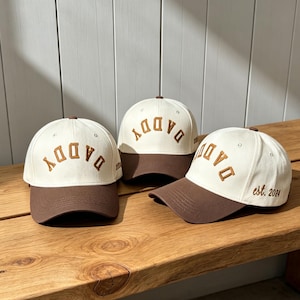 Puede incluir: Tres gorras de béisbol con corona color blanco hueso y visera marrón. Cada gorra presenta la palabra DADDY bordada en letras marrones arqueadas en el frente. Una gorra incluye el texto est. 2024 bordado en marrón en el panel lateral.