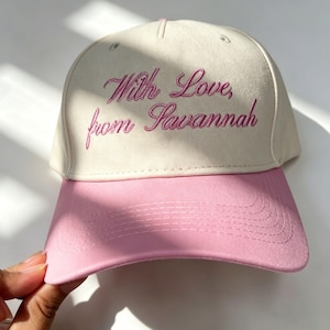 Può includere: Un cappellino da baseball color crema con visiera rosa e scritta ricamata in rosa che recita "With Love, from Savannah" in carattere corsivo. Il cappellino presenta una corona strutturata con cuciture in tinta e dettagli ad occhiello.