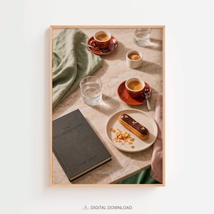 May include: A framed print featuring a black book titled The Metamorphosis by Franz Kafka, two cups of espresso on red saucers, a glass of water, a small bowl of sugar, and a chocolate eclair on a plate, all arranged on a marble surface.