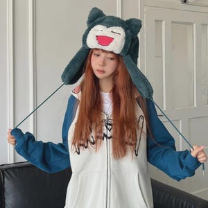 May include: A dark teal plush hat featuring a cartoon character face with a white muzzle, closed eyes, and an open mouth with a pink tongue. The hat has long ear flaps with thin dark teal strings. A white and teal zip-up hoodie is also visible.