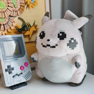 Könnte beinhalten: Ein hellgraues Plüschtier mit pixeligen schwarzen und grauen Gesichtszügen und einem runden Körper, neben einer aus grauen und weißen Bausteinen gefertigten Handheld-Spielkonsole mit dem Text Nintendo GAME BOY und zwei leuchtend rosa Knöpfen.