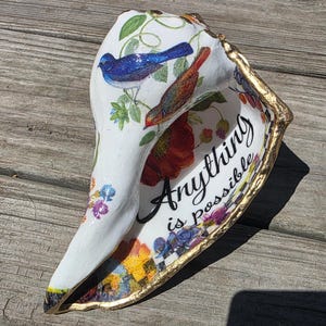 May include: A white conch shell decorated with decoupage featuring a blue bird, a brown bird, and colorful floral patterns. The interior displays the black cursive text Anything is possible. The outer edge of the shell is painted with a metallic gold trim.