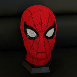 May include: A red fabric mask featuring a black web pattern across the surface. The mask includes large, almond-shaped eye lenses with a glossy black frame and a white mesh interior. The head-shaped mask is displayed on a black hexagonal base.