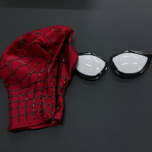 Puede incluir: Una m&aacute;scara de tela roja con un patr&oacute;n de red negro en relieve y dos lentes oculares de malla blanca separados con marcos negros. La m&aacute;scara est&aacute; hecha de un material texturizado y se muestra junto a las dos piezas oculares desmontables.