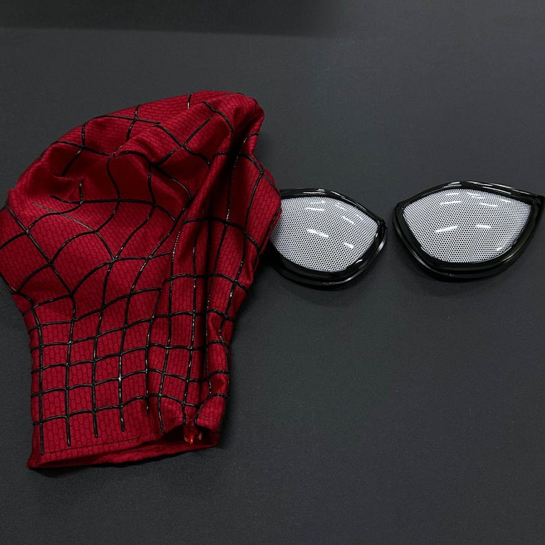 Puede incluir: Una m&aacute;scara de tela roja con un patr&oacute;n de red negro en relieve y dos lentes oculares de malla blanca separados con marcos negros. La m&aacute;scara est&aacute; hecha de un material texturizado y se muestra junto a las dos piezas oculares desmontables.