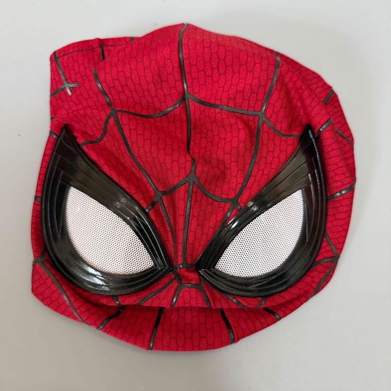 Puede incluir: Una m&aacute;scara de tela roja con un patr&oacute;n de telara&ntilde;a negra en la superficie. La m&aacute;scara incluye dos lentes grandes en forma de almendra con marcos negros e inserciones de malla blanca, dise&ntilde;adas para cubrir la parte superior de la cabeza.