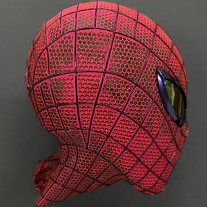 May include: A red mask with a black honeycomb texture pattern and black web lines. The mask features a single visible eye lens with a dark metallic frame and a reflective yellow-tinted surface, covering the upper portion of a head-shaped form.