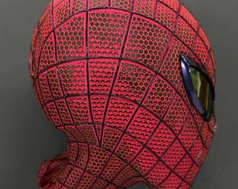 Puede incluir: Una máscara roja con un patrón de textura de panal negro y líneas de telaraña negras. La máscara presenta una lente ocular visible con un marco metálico oscuro y una superficie reflectante con un tinte amarillo.