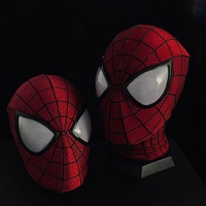 Pode incluir: Duas máscaras de tecido vermelho com um padrão de teia preto e grandes lentes oculares de malha branca. Uma máscara é exibida num suporte preto, enquanto a outra está à esquerda. Ambas têm uma superfície texturizada e bordas pretas.
