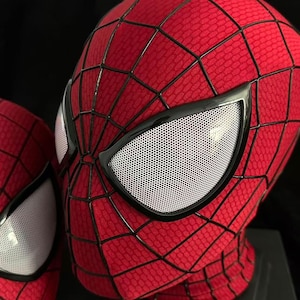 Puede incluir: Una m&aacute;scara de tela roja con un patr&oacute;n de telara&ntilde;a negro y lentes oculares grandes, blancos y cubiertos de malla. La m&aacute;scara presenta una superficie texturizada y un borde negro distintivo alrededor de cada ojo con un dise&ntilde;o de rejilla.