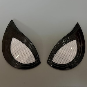 Puede incluir: Un par de lentes oculares estilizadas en forma de almendra, con un marco negro brillante y un centro de malla blanca. El dise&ntilde;o es afilado en las esquinas exteriores, creando una apariencia angular y n&iacute;tida para una m&aacute;scara o accesorio.