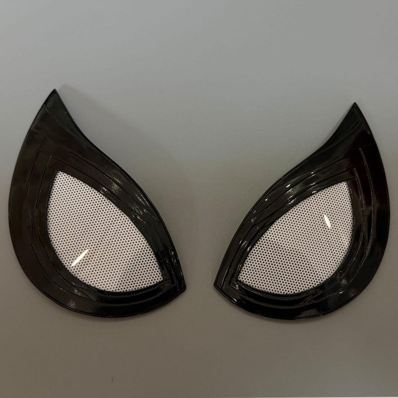 Puede incluir: Un par de lentes oculares estilizadas en forma de almendra, con un marco negro brillante y un centro de malla blanca. El dise&ntilde;o es afilado en las esquinas exteriores, creando una apariencia angular y n&iacute;tida para una m&aacute;scara o accesorio.