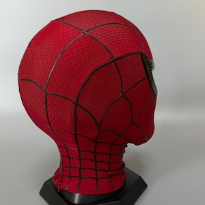 Puede incluir: Una m&aacute;scara de tela roja con un patr&oacute;n de red negro en la superficie y lentes oculares oscuros y reflectantes. La m&aacute;scara se muestra sobre un soporte negro, exhibiendo un material texturizado con un dise&ntilde;o de rejilla detallado.