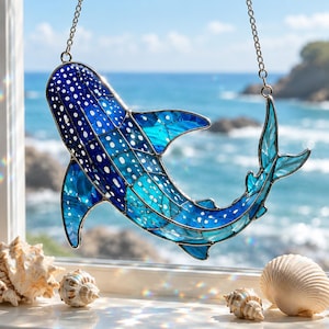 Pode incluir: Um apanhador de sol de vitral em forma de tubarão-baleia, apresentando segmentos de vidro azul profundo e turquesa com padrões de pontos brancos. A peça está suspensa por uma corrente de metal prateado, exibindo linhas de chumbo detalhadas.