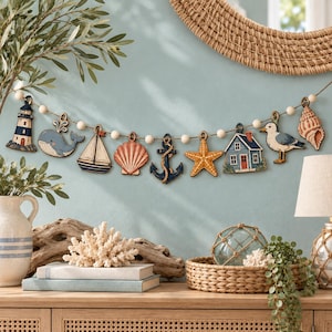 May include: A nautical-themed garland featuring wooden cutouts of a lighthouse, whale, sailboat, seashell, anchor, starfish, house, seagull, and conch shell, all strung together with white wooden beads on a natural twine cord.