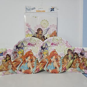 Może przedstawiać: Zestaw artykułów imprezowych Winx Club zawierający dwa okrągłe papierowe talerze i trzy opakowania papierowych serwetek. Przedmioty zawierają kolorowe ilustracje wróżek z pomarańczowymi, blond i ciemnymi włosami na kwiatowym tle.