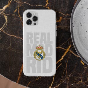 May include: A clear smartphone case featuring the text REAL MADRID in large light gray letters and the official Real Madrid football club crest in the center, displaying a gold crown, blue and yellow details, and a blue diagonal stripe.
