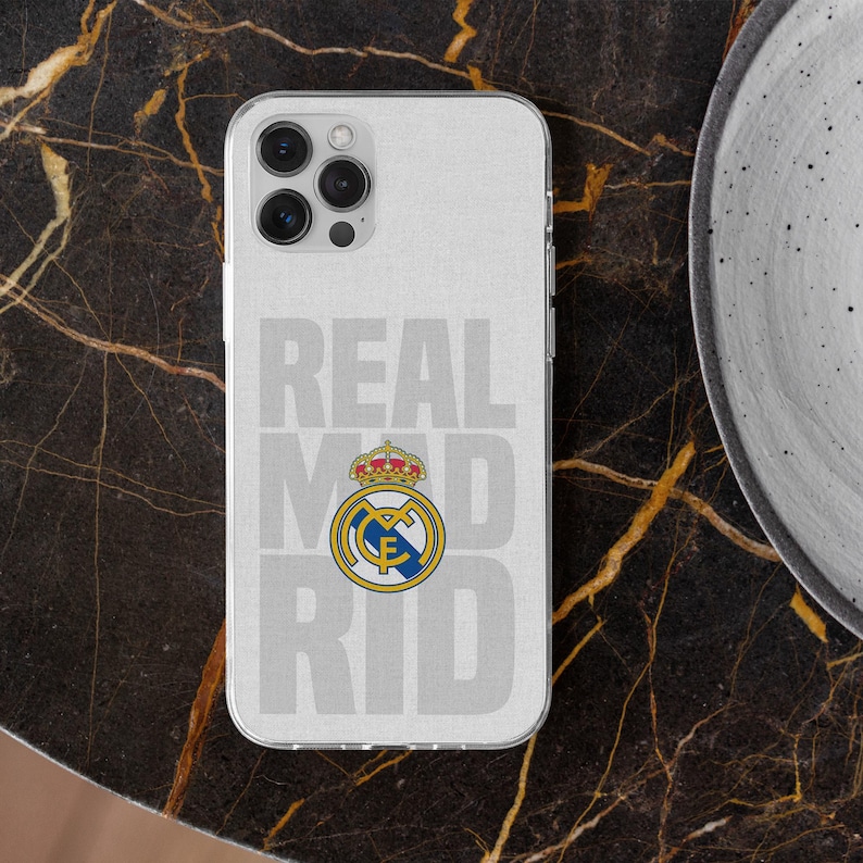 May include: A clear smartphone case featuring the text REAL MADRID in large light gray letters and the official Real Madrid football club crest in the center, displaying a gold crown, blue and yellow details, and a blue diagonal stripe.