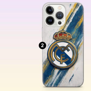 May include: A clear smartphone case featuring a Real Madrid crest with a gold crown and blue and gold diagonal brushstroke accents on a white background. The design includes a prominent number two in a black circle on the left side of the case.