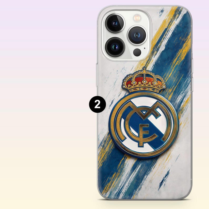 May include: A clear smartphone case featuring a Real Madrid crest with a gold crown and blue and gold diagonal brushstroke accents on a white background. The design includes a prominent number two in a black circle on the left side of the case.
