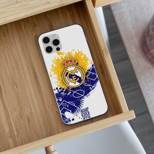 May include: A white smartphone case featuring a Real Madrid soccer club logo with a crown, set against a yellow and blue graphic design with soccer field markings and a silhouette of a cheering crowd, including a small secondary club emblem.