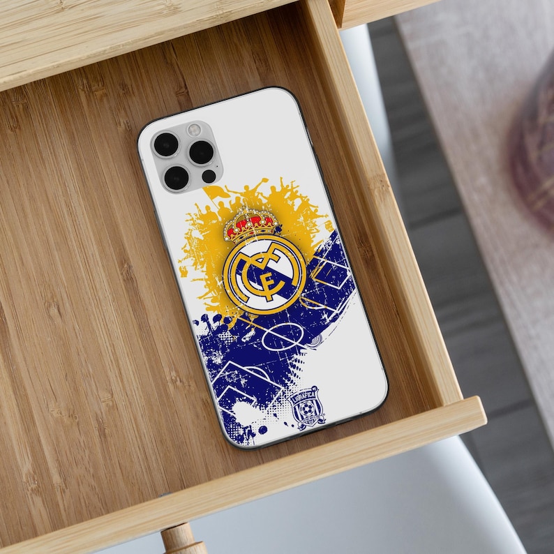 May include: A white smartphone case featuring a Real Madrid soccer club logo with a crown, set against a yellow and blue graphic design with soccer field markings and a silhouette of a cheering crowd, including a small secondary club emblem.