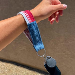 Puede incluir: Un llavero de muñeca de tela con una sección de mezclilla azul, una sección con patrón de cuadros rojos y blancos y un forro de tela blanca. La pulsera está unida a una anilla de metal plateado que sujeta un mando de llave de plástico negro.